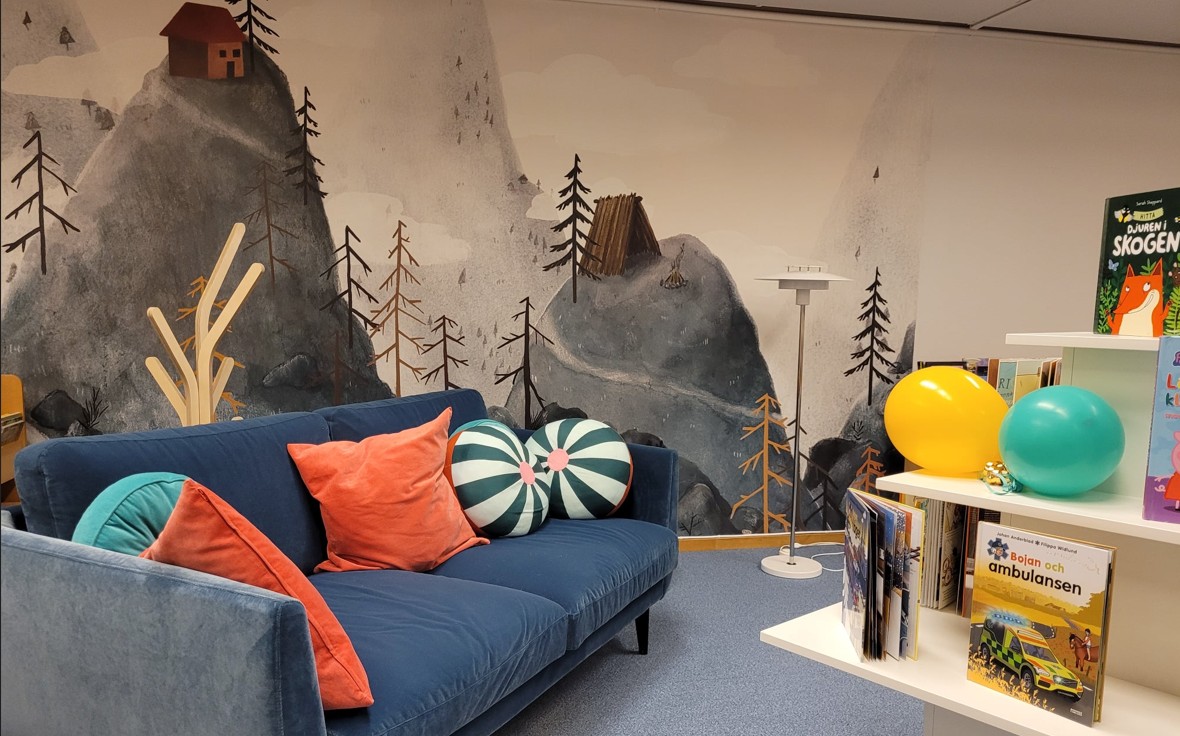 Biblioteksmiljö med blå sammetssoffa, färglada kuddar mot en vägg med naturillustration.